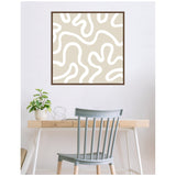 Square framed canvas with beige background and abstract white curvy lines, hanging above a light wood desk with a small potted plant and wire sculpture, facing a gray chair.
