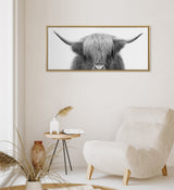 Black and white photograph of a Highland cow with long shaggy hair covering its eyes, framed in gold and hung on a white wall above a beige armchair and wooden side table.