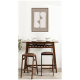 Minimalist framed artwork with two abstract organic white shapes on beige background above a narrow wooden bar table holding white dishes, flanked by two wooden stools on a light floor