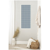 Tall rectangular pinboard with light blue plaid fabric and white grid lines, framed in white, hanging on a light gray wall above a wicker basket and decorative vase.