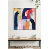 Abstract framed canvas with broad curved strokes in navy, red, yellow, pink, and white, hanging above a wooden console with books, a mug, and a white vase with greenery.