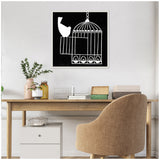 Black and white linocut art showing a bird perched on an open birdcage door, framed and hung above a wooden desk with a beige upholstered chair.
