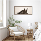 Framed canvas print showing close-up of black horse ears and mane against a soft beige background, hanging above a wicker chair and rustic wooden bedside table.