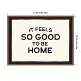 Framed canvas print with black text saying 'It feels so good to be home' on a white background, bordered by a black inner frame and a brown outer frame measuring 24 by 18 inches.