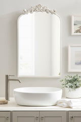 Rectangular wall mirror with an arched top and ornate cream-colored floral carvings above, reflecting a simple light-colored wall in a bathroom setting.