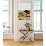 Abstract landscape painting with muted greens, browns, and purples in a simple white frame, hung on a beige wall between two tall windows above a gold and wood bar cart.