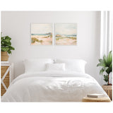 Two square framed canvases above a white bed showing soft, pastel beach landscapes with sand dunes, grasses, and calm water under a pale sky.