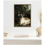 Framed canvas showing a reindeer lying peacefully under a Christmas tree with hanging red ornaments in a dimly lit setting.