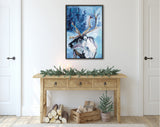 Framed canvas painting of a reindeer with large antlers in snowy blue landscape, hung above a rustic wooden console table decorated with pine garland and candles.