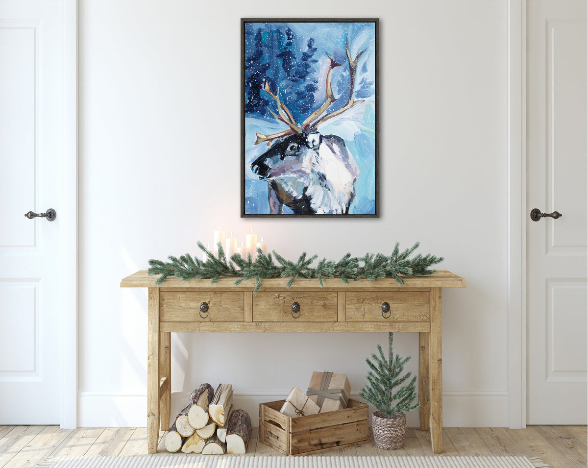 Framed canvas painting of a reindeer with large antlers in snowy blue landscape, hung above a rustic wooden console table decorated with pine garland and candles.