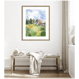 Watercolor painting of a rustic barn in a green field under a partly cloudy blue sky, framed and hung on a white wall above a wooden bench and light-colored drapery.