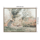 Framed canvas print showing a cheetah lying on grass with a blurred background of trees and bushes in soft earthy tones.