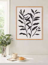 Black botanical silhouette print with intertwined leaves in a light wooden frame, hanging above a white table with a cup, croissant, and glass vase of greenery near a window.