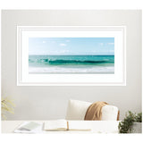 Framed panoramic photo of a turquoise ocean wave gently breaking under a blue sky with scattered clouds, displayed above a white chair with a tan throw.