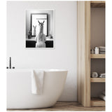 Black and white framed canvas showing a llama facing a mirror with its reflection and a small potted plant on each side, mounted on a white textured wall above a modern white bathtub.