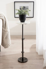Small round white marble top table with adjustable black metal stand holding a black pot with green leafy plant, placed on a light wooden floor near a beige knitted blanket and white curtains.