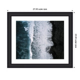 Aerial view of ocean waves crashing with white foam onto a textured black sand beach, framed in a sleek black frame with white matting.