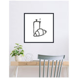 Minimalist black line drawing of a steaming cup and croissant on white canvas framed in black, hung above a light wood desk with a potted plant and a gray chair.
