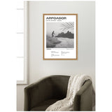 Framed black and white wall art showing a surfer riding a wave near Arpoador, Rio de Janeiro, with a palm leaf shadow in the top right corner, hung above a dark olive chair with a knitted throw.