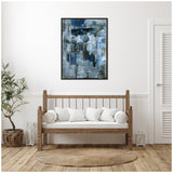 Abstract painting with blue, gray, and white hues in a black frame hangs above a wooden bench with white cushions, flanked by a potted plant and a lantern, in a minimalist room.