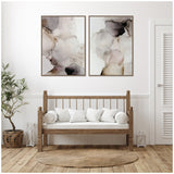 Two framed abstract canvas prints with soft smoke-like patterns in shades of gray, beige, and brown above a wooden bench with white cushions in a minimalist room.