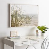 Framed canvas of beach grass and sand dunes with muted green and beige tones, hanging above a white desk featuring a plant, pencils, and a 'GOOD VIBES' sign.