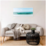 Light gray modern sofa with a small dog lying on it, accompanied by a gray throw and round beige pillow, in a room with a black wicker coffee table and framed ocean wave photograph above.