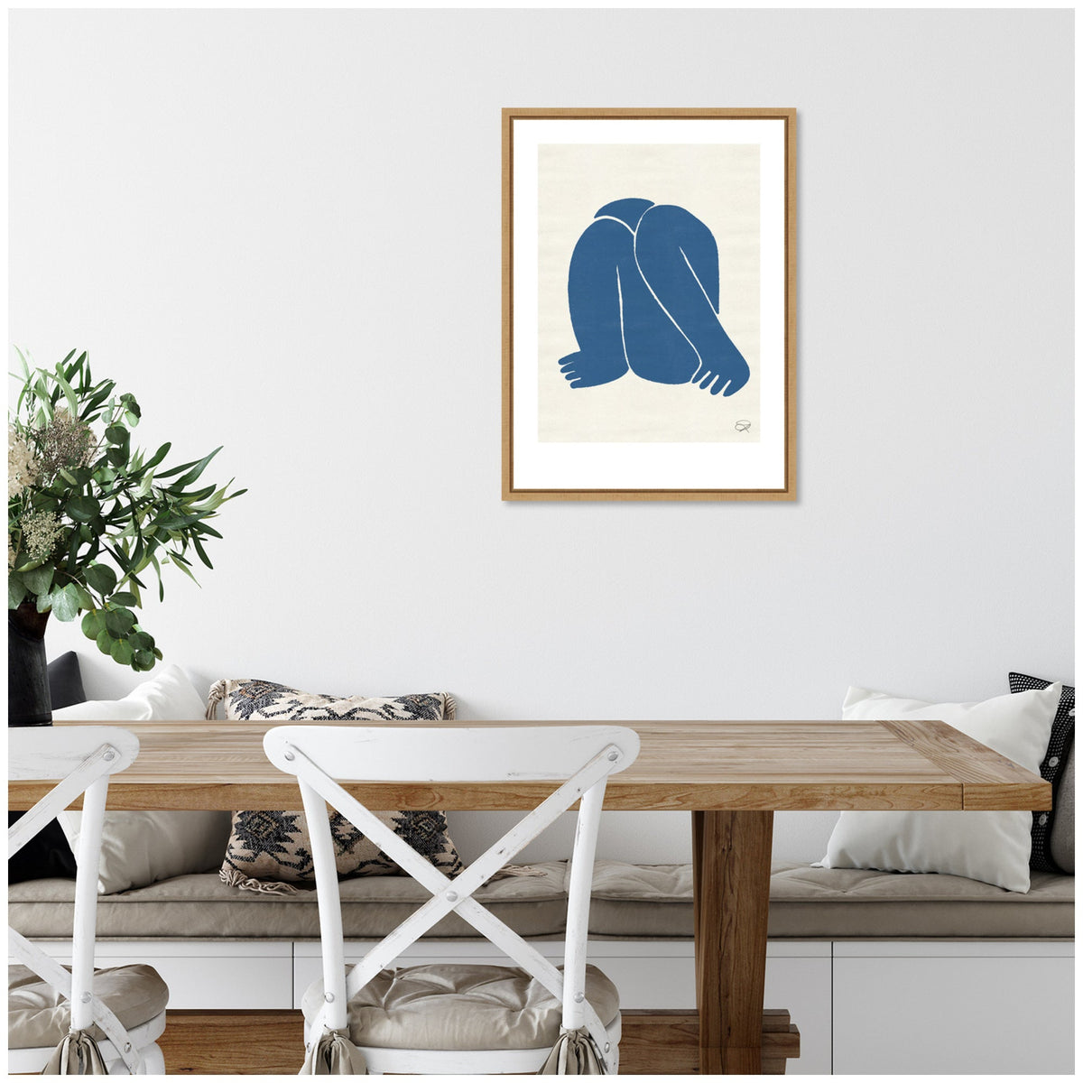 Framed canvas art showing a minimalist blue figure with folded legs and arms, hanging on a white wall above a wooden dining table with white chairs and cushions.