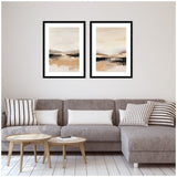 Two framed abstract landscape prints in beige, black, and muted peach tones hang above a gray sectional sofa with striped and solid pillows in a minimalist living room.