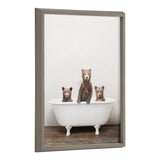 Three brown bear cubs standing and sitting inside a white vintage clawfoot bathtub on a wooden floor against a plain light wall, framed in grey wood.