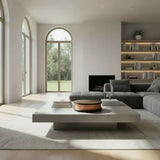 Minimalist living room with large arched windows, a low rectangular coffee table in light stone, and a gray sectional sofa facing built-in illuminated bookshelves.