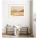 Framed abstract landscape with soft pastel folds of peach, green, brown, and blue above a wooden bench with a book and draped fabric in a bright minimal room.