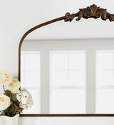 Top corner of a traditional arch mirror with a decorative bronze-colored applique and visible reflections of white window panes behind cream-colored roses in a white vase.