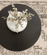 Top-down view of a round black wood table with a white ceramic vase holding slender branches with small white flowers, sitting on a beige rug with black geometric patterns.