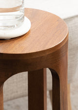 Close-up of a round, smooth-edged wooden table top with tapered legs, holding a clear glass of water on a white coaster, beside a gray fabric surface.