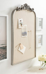 Arched pinboard with silver ornamental frame, covered in beige fabric, displaying photos and jewelry, mounted on a white wall with framed pictures nearby.