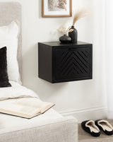 Matte black floating side table with a chevron-patterned door, topped with two black vases holding dried pampas grass and cotton stems, mounted beside a bed.