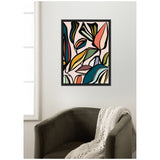 Framed abstract botanical wall art with multicolored leaves in shades of pink, green, blue, and terracotta on a pale pink background, hung above a dark olive armchair with a cream knit throw.