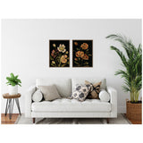 Two framed floral canvas artworks with warm-toned flowers on black backgrounds hung above a white sofa with patterned and solid cushions, flanked by green potted plants.