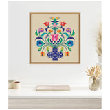 Square canvas wall art featuring a symmetrical bouquet of colorful stylized flowers in a blue vase on a beige background, framed in light wood and hung above a white surface with dried grasses.