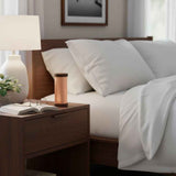 Copper-colored cylindrical Qi-Shield by Waveguard device on dark wooden bedside table with white lamp, open book, and eyeglasses next to a bed with white pillows and sheets.