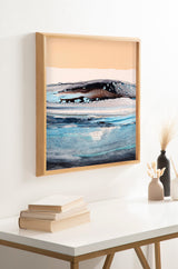 Abstract framed art with fluid patterns in blues, black, and white beneath a tan background, displayed on a white wall above a table with books and vases.