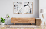 Two framed abstract canvas art pieces with black curved lines and gray, white, beige tones hanging above a light wooden sideboard in a minimalist room with plants and a woven basket.