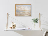 Soft pastel beach landscape print with gentle waves and cloud-filled sky in light wood frame hanging above a minimalist white hanging shelf with gold geometric decor and a glass vase with greenery.