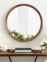 Round wall mirror with a smooth medium-brown wooden frame reflects a soft window and green plant, mounted above a wooden shelf with books and white flowers.