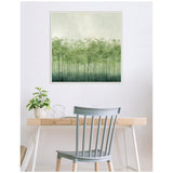 Square canvas with an abstract forest of delicate green tree silhouettes fading into a soft misty background, hung above a light wood desk with a gray chair and plant.