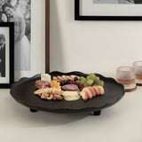 Black round tray with scalloped edges holding an assortment of cheeses, nuts, sliced apples, grapes, and cured meats, set on a white surface near framed photos and textured glasses.