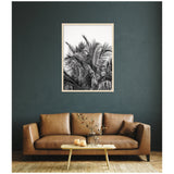 Black and white framed photo of palm tree leaves hangs centered on a dark green textured wall above a brown leather sofa with a single pillow and a wooden coffee table with glass vase and two candles