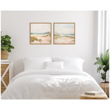 Two framed square canvas paintings above a white bed with cushions, featuring soft pastel landscapes of sandy dunes and a cloudy sky.