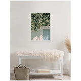 Framed canvas print of white jasmine blossoms cascading over a pastel pink wall with a closed pale blue door, hung above a white bench with a furry throw and beige bag.